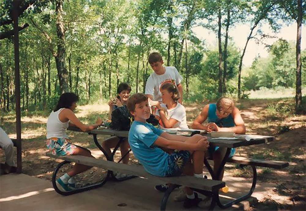 A group of kids sit and stand around a picnic table in a wooded outdoor area on a sunny day, some talking and laughing while others write or draw. The scene is relaxed and summery, with trees in the background.