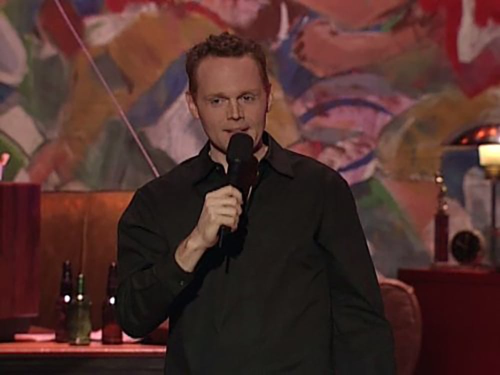 A man in a black shirt stands on stage holding a microphone, with colorful abstract art in the background and bottles and a lamp visible on a table behind him.