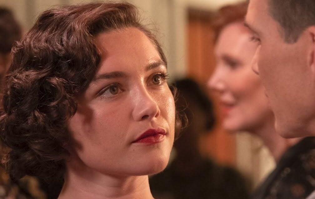 A woman with short, wavy brown hair and red lipstick looks intently at a man in a formal setting, with blurred people in the background.