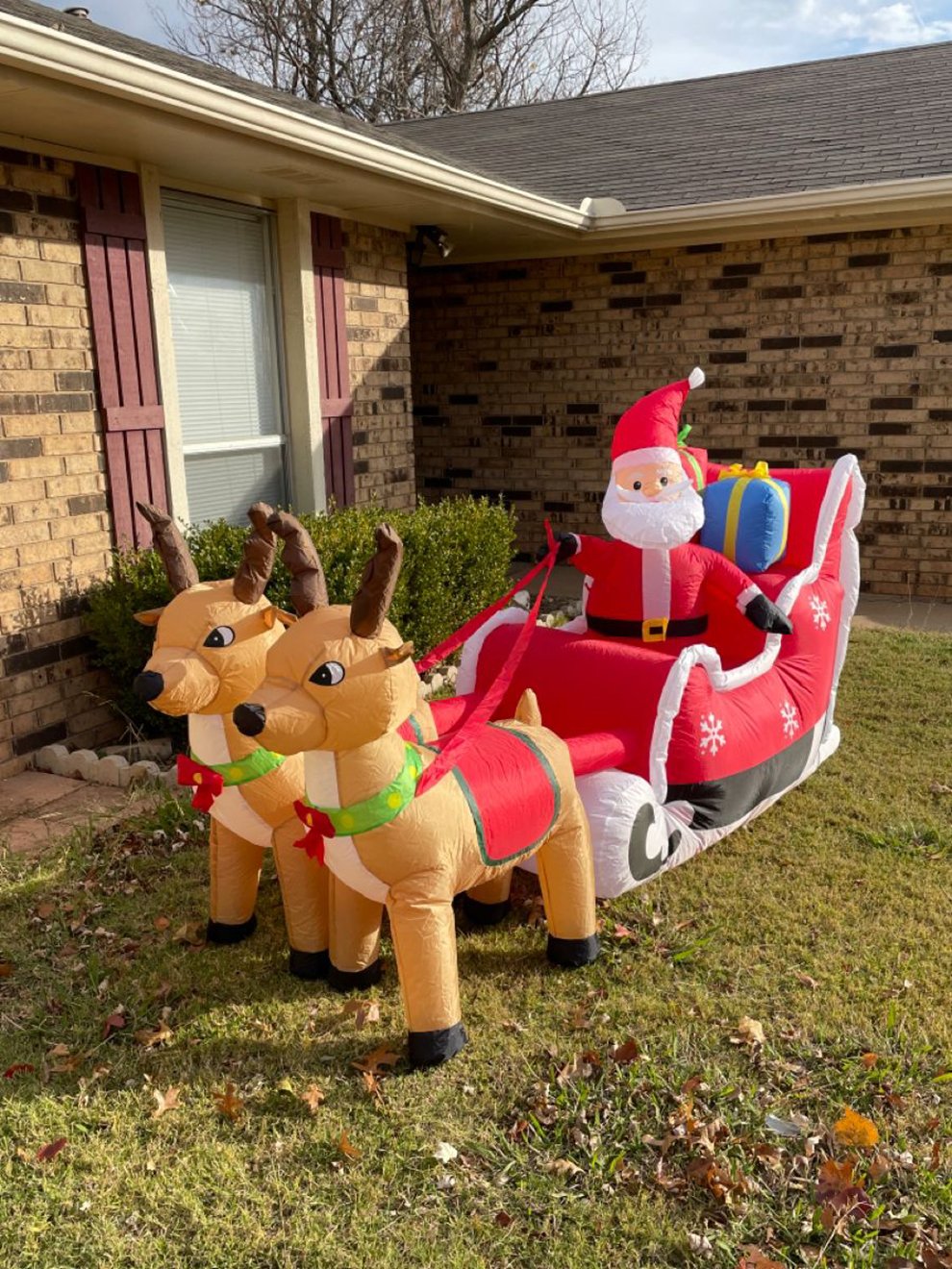 An inflatable Santa Claus sits in a red sleigh pulled by two inflatable reindeer, displayed on the lawn outside a brick house with red shutters.