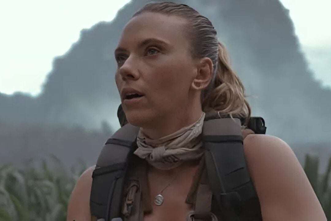 A woman with light hair tied back, wearing a scarf around her neck and a backpack, stands outdoors with a determined expression. Misty mountains and greenery are visible in the background.