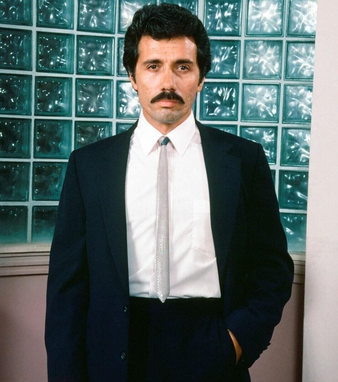 A man with dark hair and a mustache stands in front of glass block windows, wearing a dark suit, white shirt, and light-colored tie, with one hand in his pocket and a neutral expression.