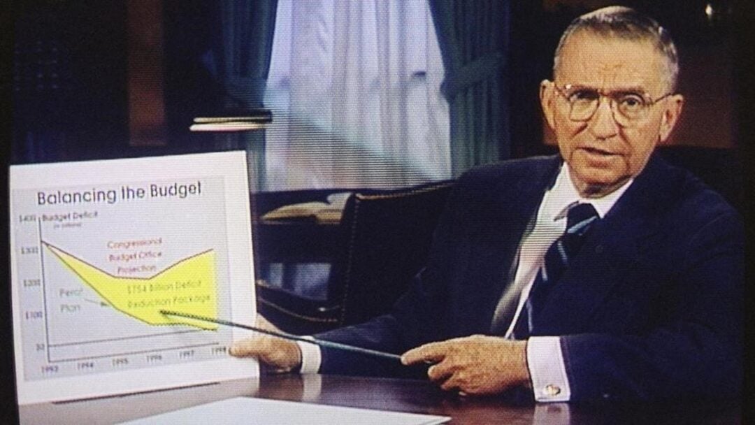 An older man in a suit sits at a desk, pointing at a line graph titled “Balancing the Budget” with a pointer. The graph compares projected deficits, and a curtain is visible in the background.