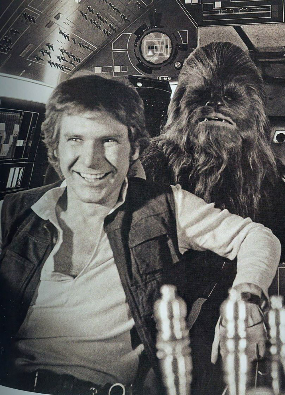 A man with a vest and open shirt smiles while seated at a spacecraft control panel, with a tall, furry creature standing behind him in a sci-fi setting.