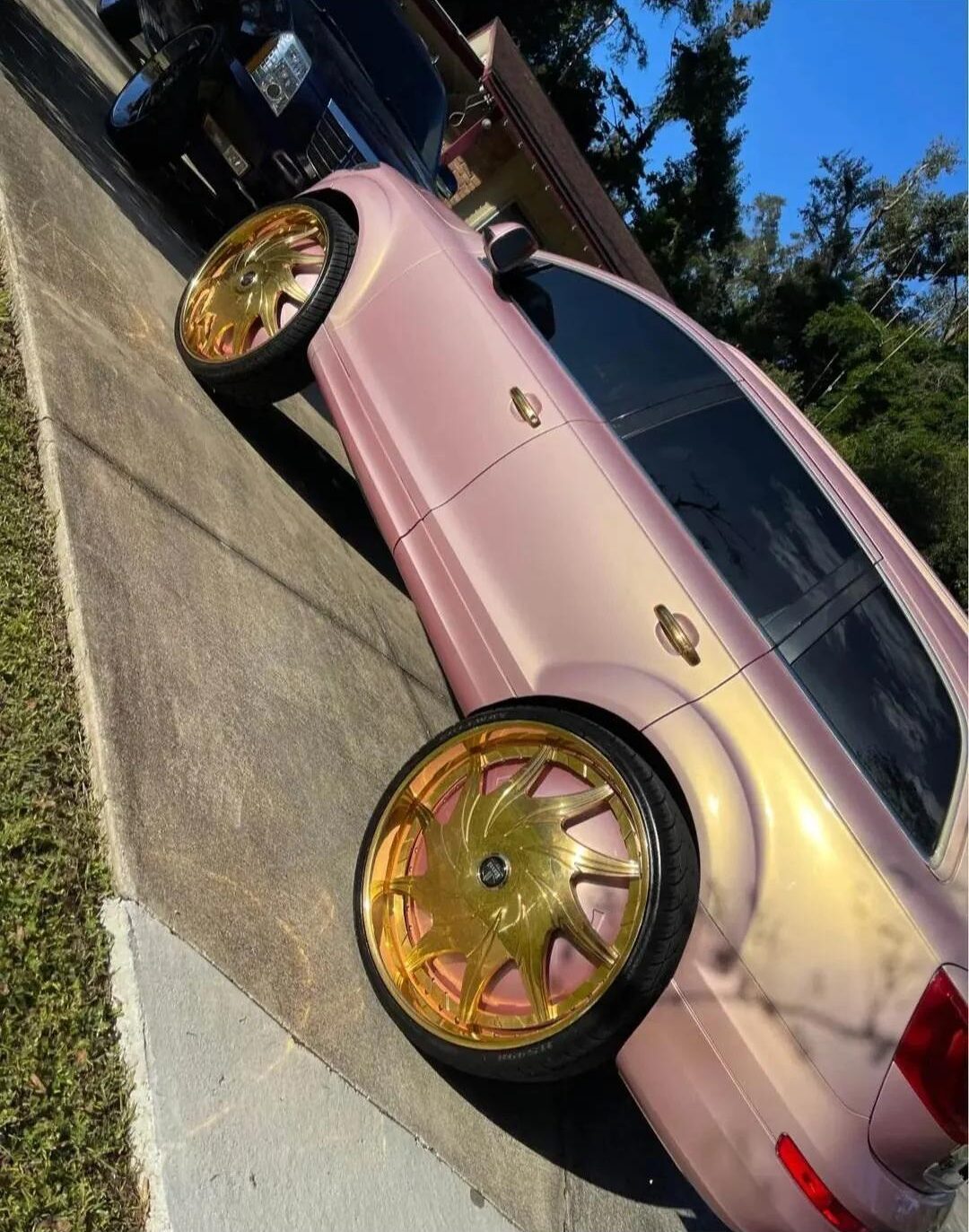 A pink SUV with large, shiny gold rims is parked on a driveway next to a black vehicle, with trees and a house visible in the background.