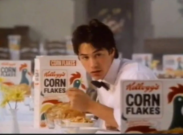 A young man in a white shirt and black bow tie eats cereal at a table lined with multiple Kellogg’s Corn Flakes boxes and vases of yellow flowers. He looks up from his bowl toward the camera.