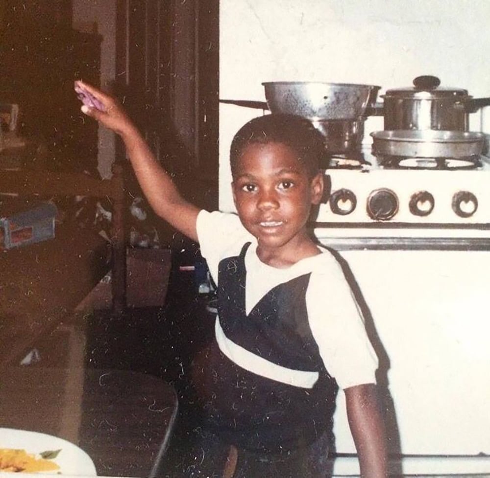A young boy smiles while standing in a kitchen, raising one arm. Behind him are a stove with pots and a pan, and various kitchen items. He wears a white shirt under dark overalls.