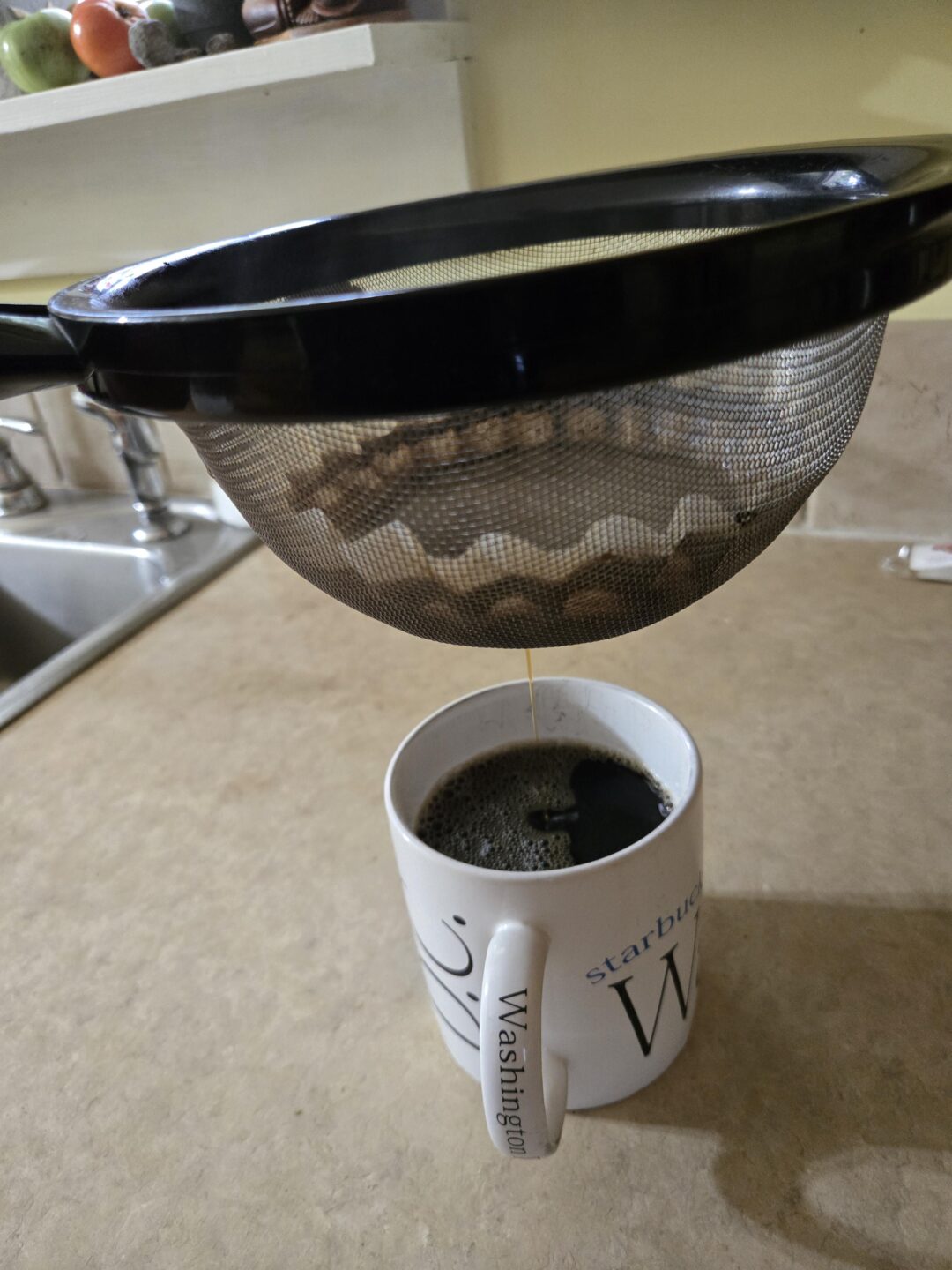 A metal strainer with a coffee filter is held above a white mug, filtering coffee into the cup on a kitchen countertop. A sink and fruit on a shelf are visible in the background.