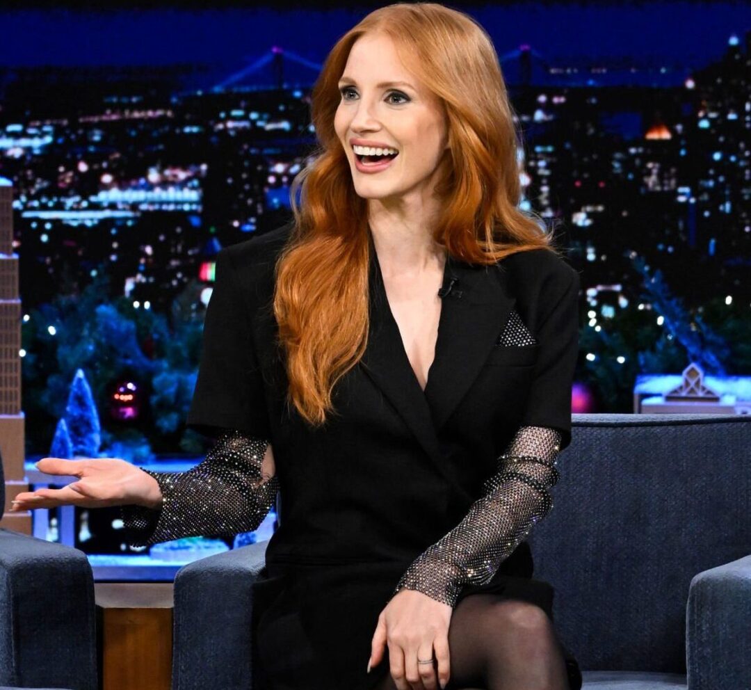 A woman with long red hair, wearing a black outfit with sparkly sleeves, sits on a talk show set at night, smiling and gesturing with her hand. The city skyline is visible in the background.