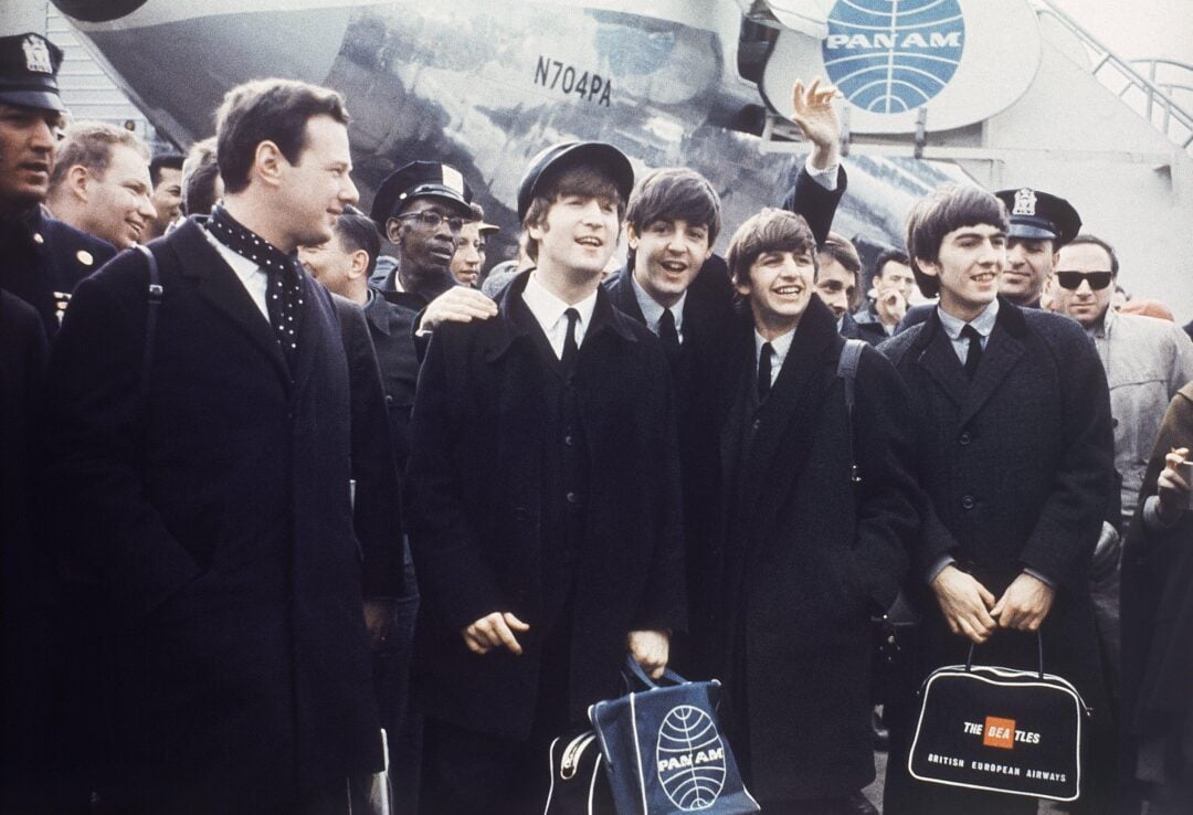 Four young men in suits and ties stand smiling and waving in front of a Pan Am airplane, surrounded by a crowd, police officers, and holding travel bags. The scene appears to be at an airport in the 1960s.