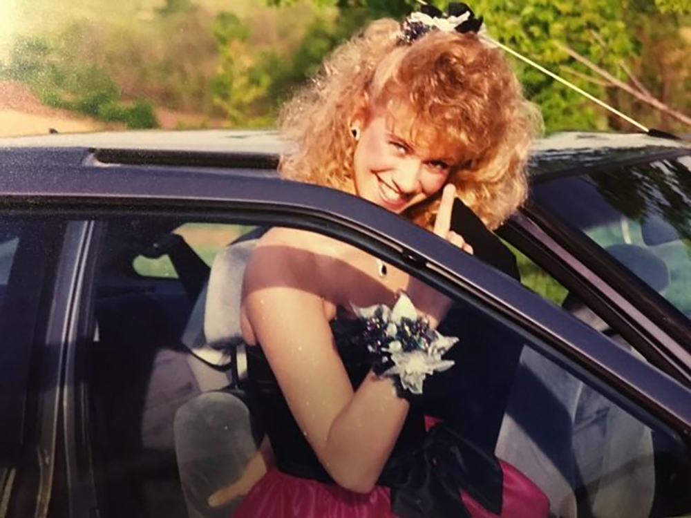 A young woman dressed in a formal gown with a corsage on her wrist smiles while standing next to an open car door, holding up her index finger, with greenery in the background.