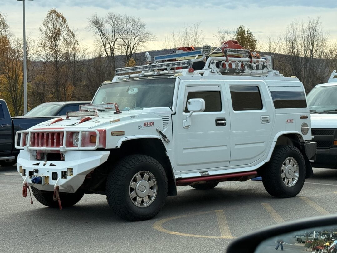 A white Hummer H2 is parked in a lot. The vehicle is heavily modified with extra lights, racks, pipes, and gear on the roof and hood. Trees and hills are visible in the background.