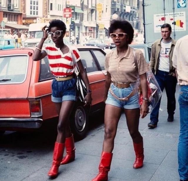 Two women wearing red boots, denim shorts, and sunglasses walk confidently down a city street. One wears a red-striped shirt, the other a beige polo. Cars and pedestrians are visible in the urban background.