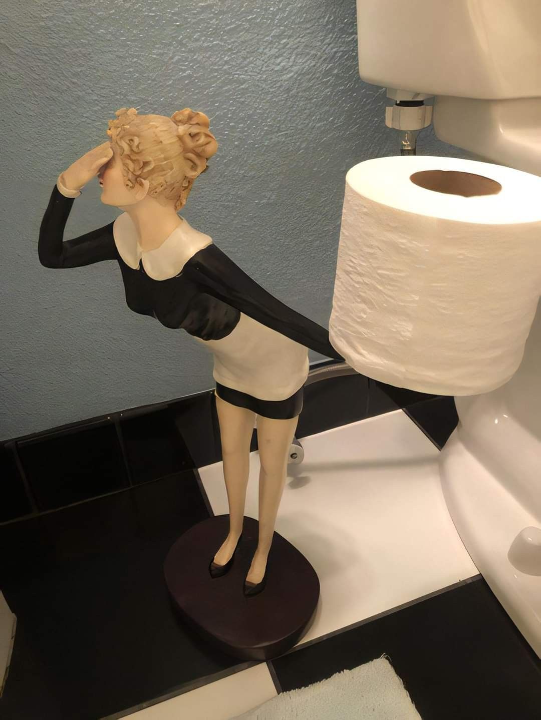 A figurine of a woman in a black and white dress is positioned next to a toilet, holding a roll of toilet paper behind her back as if serving it, with one hand raised to her forehead in a searching gesture.