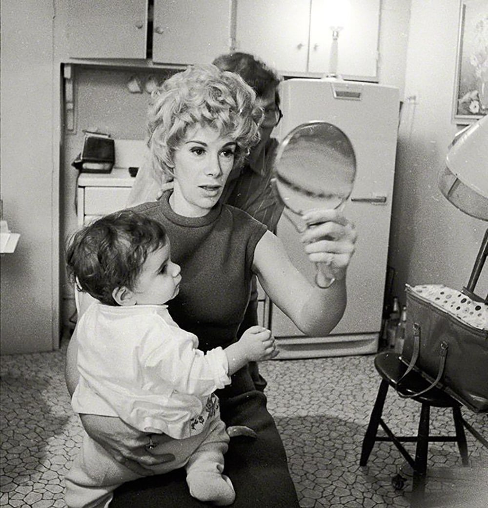 A woman with curly hair holds a baby on her lap, looking into a handheld mirror in a vintage kitchen. A man is partially visible behind her. A large purse and a hooded hair dryer are nearby.