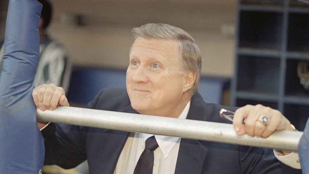 A man in a suit and tie smiles while leaning on a metal railing, holding it with both hands. He has short, light hair and is wearing a large ring on his right hand. The background appears to be a sports setting.