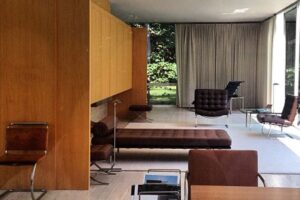 A modern living room with floor-to-ceiling windows, wood paneling, minimalist furniture, and a view of greenery outside. The space features lounge chairs, a desk, and neutral-colored decor.