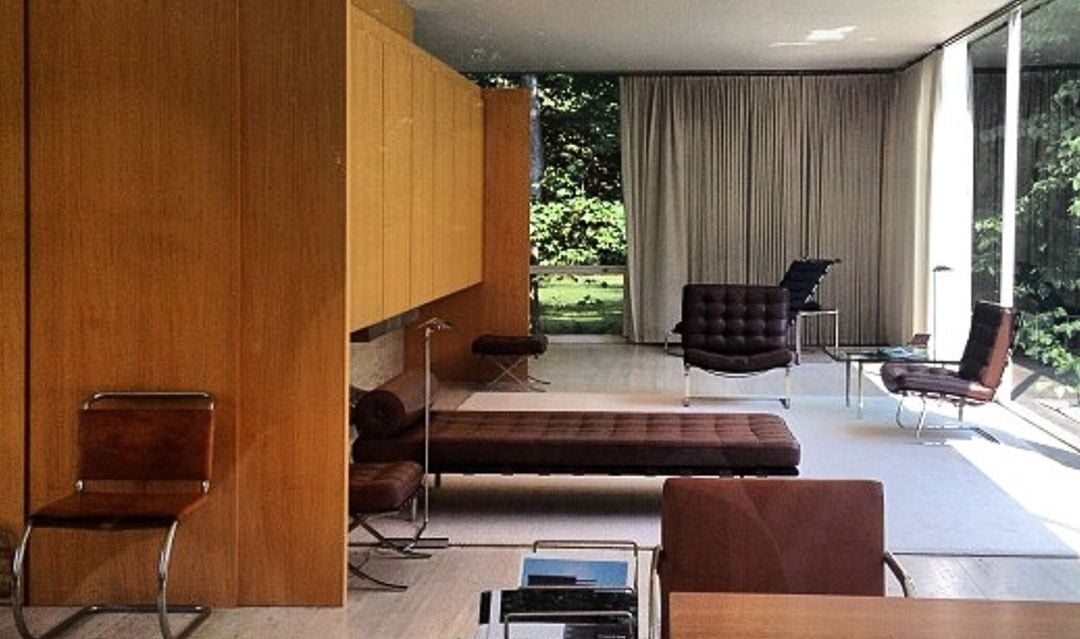 A modern living room with floor-to-ceiling windows, wood paneling, minimalist furniture, and a view of greenery outside. The space features lounge chairs, a desk, and neutral-colored decor.