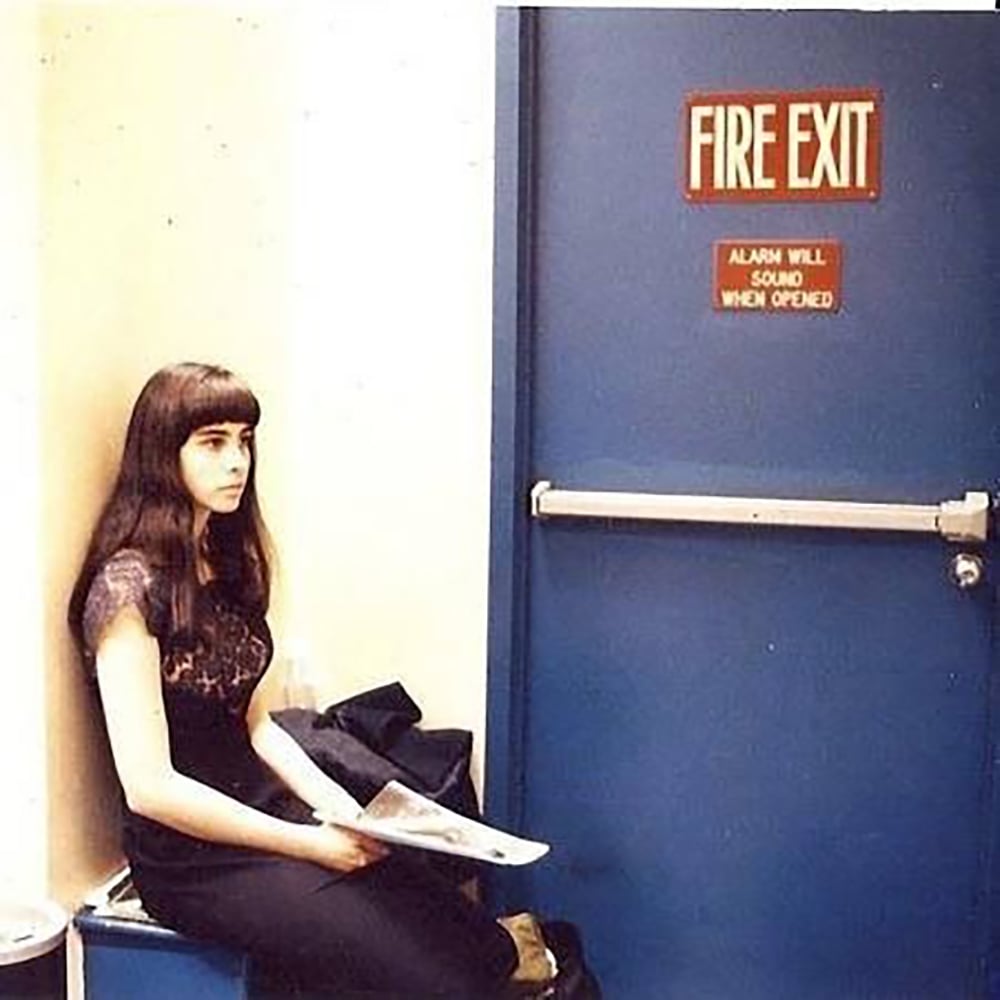 A woman with long dark hair sits on a bench near a blue fire exit door, holding papers in her lap. The sign above the door reads, "FIRE EXIT. ALARM WILL SOUND WHEN OPENED.