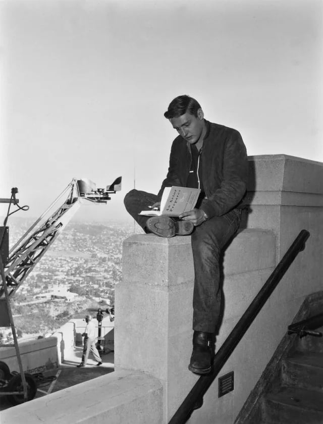 A man sits casually on a stone railing, reading a script, with film equipment and crew visible in the background and a cityscape below.