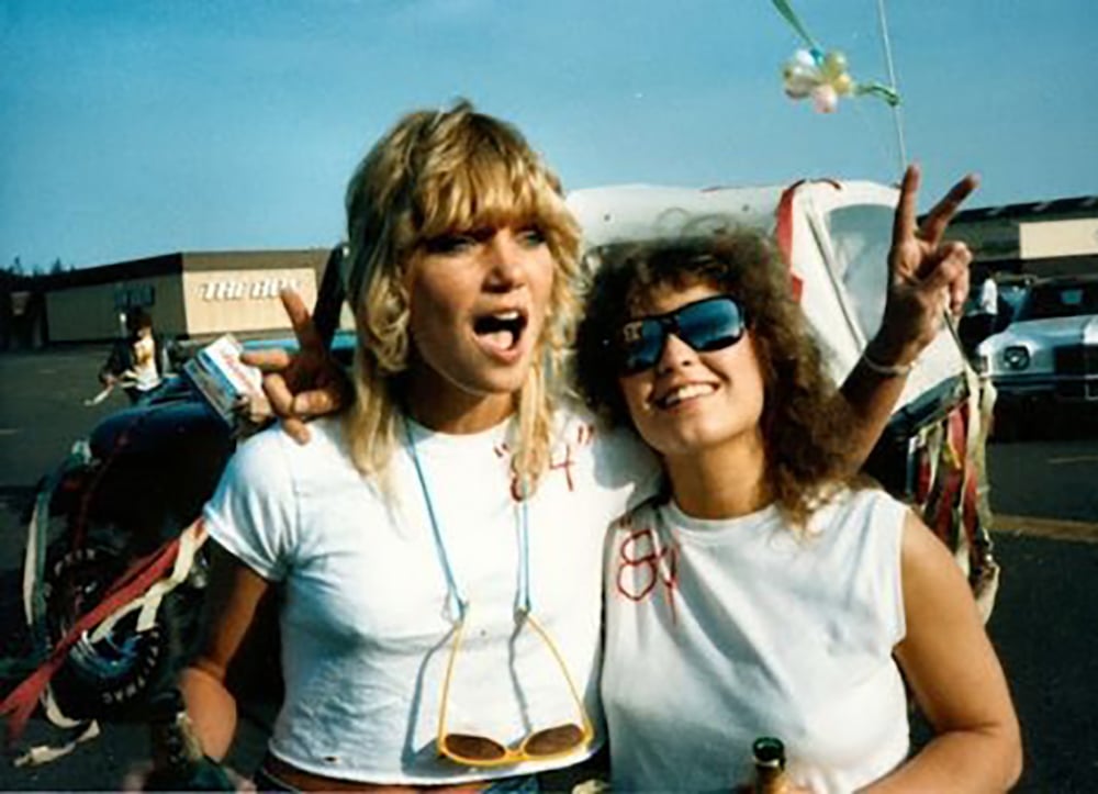 Two women wearing white t-shirts and sunglasses stand arm in arm, smiling and making peace signs at an outdoor event. Colorful streamers decorate a car in the background on a sunny day.