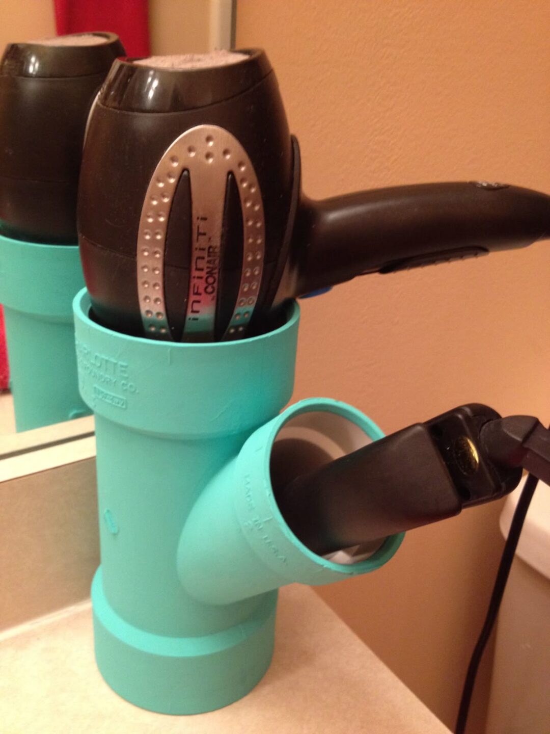 A turquoise PVC pipe organizer holds a hair dryer and a hair straightener on a bathroom counter, with a mirror reflecting part of the setup.