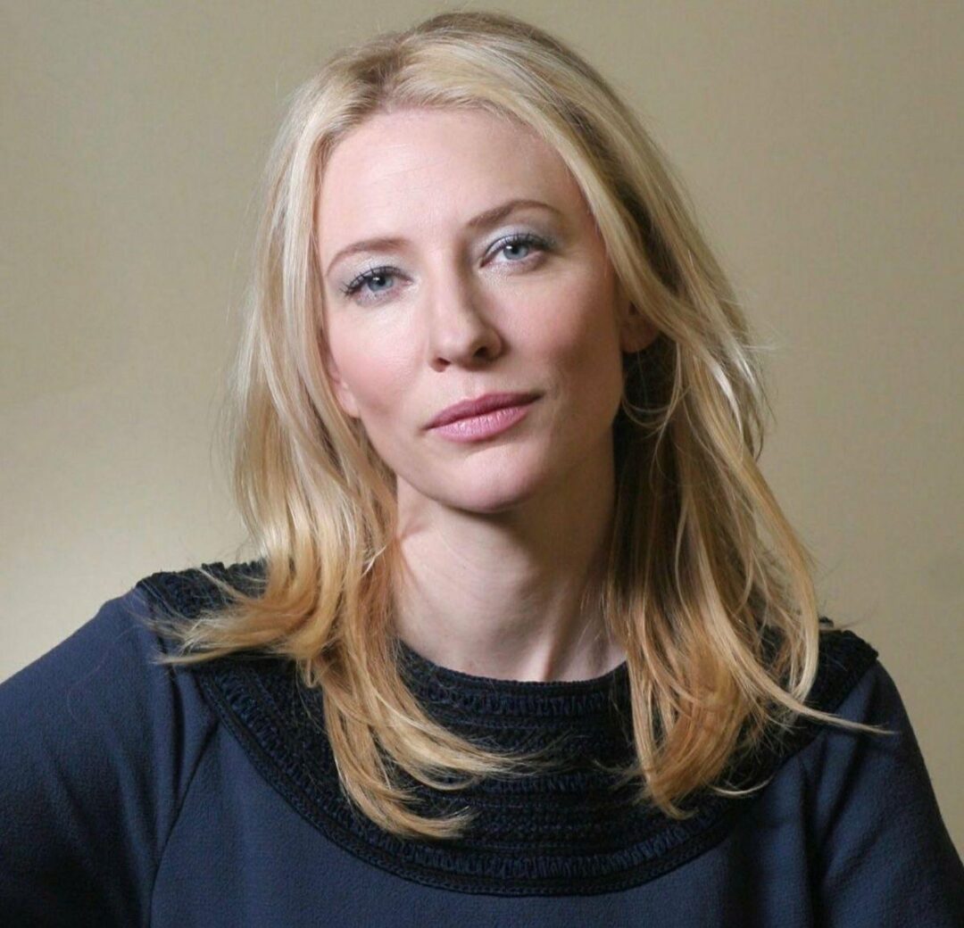 A woman with long blonde hair and light skin, wearing a dark top, sits in front of a plain beige background and looks directly at the camera with a neutral expression.