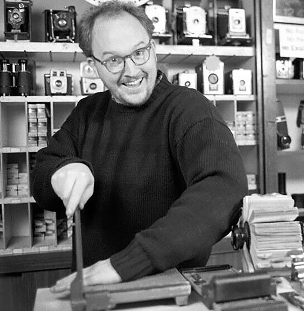 A smiling man in glasses and a dark sweater uses a paper cutter in a shop filled with vintage cameras and photography equipment.