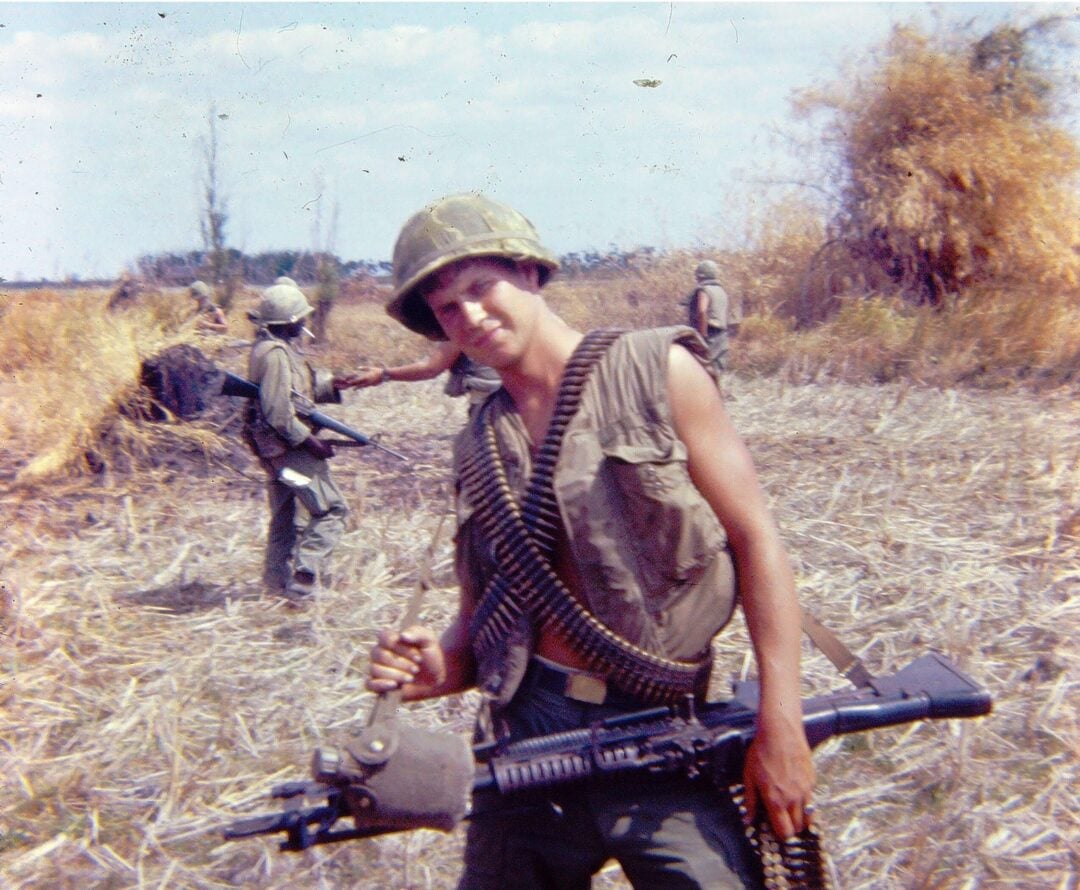 A young soldier in a helmet stands in a dry, grassy field holding a machine gun, with a bandolier of ammunition across his chest. Several other soldiers are visible in the background.