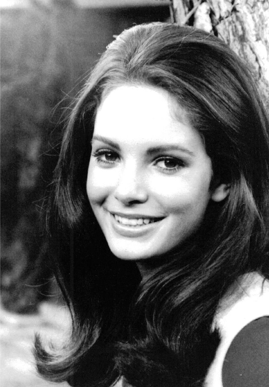 A young woman with long dark hair smiles at the camera. She is outdoors, standing near a tree, and is wearing a light-colored top. The image is in black and white.