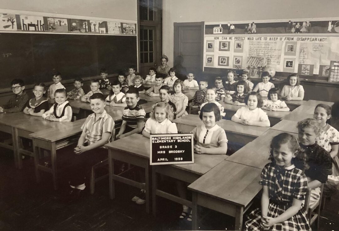 A black-and-white photo of a classroom from 1959 shows young students sitting at desks, smiling at the camera, with their teacher at the back. A sign in front reads, "Baltimore Highlands Elementary School, Grade 3, Mrs. Mockbee, April 1959.