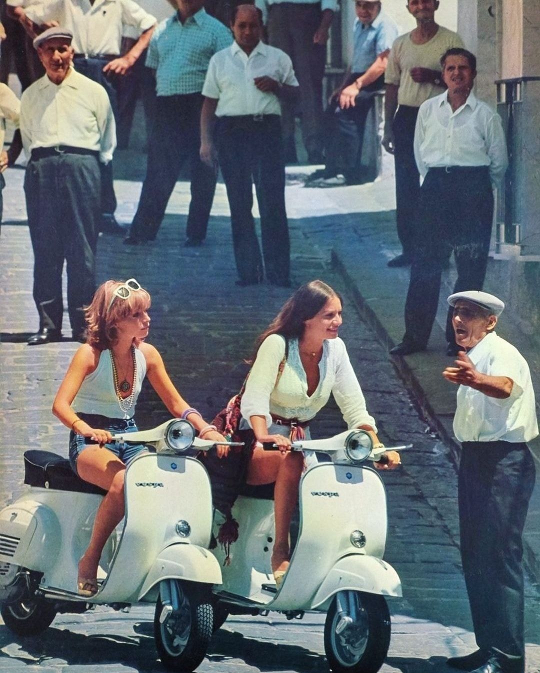 Two young women ride white Vespa scooters on a cobblestone street, smiling as they pass an older man gesturing towards them. Several men in shirts and hats stand and watch in the background.