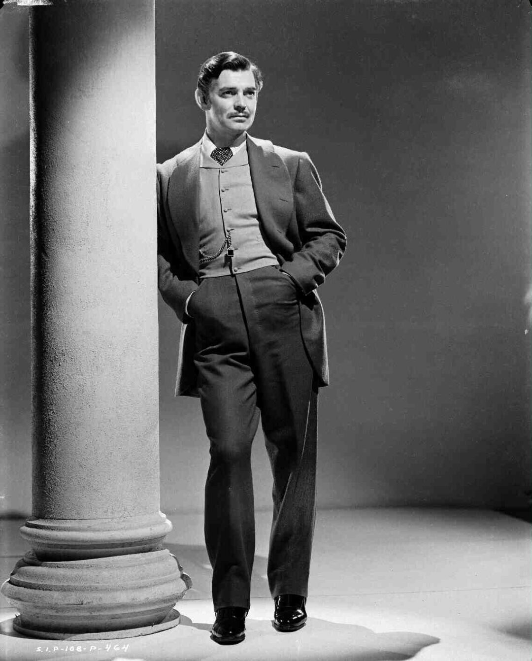 A man in a vintage suit stands with his hands in his pockets, leaning casually against a tall column. He has neatly styled hair and a mustache, looking confident in a classic studio setting with dramatic lighting.