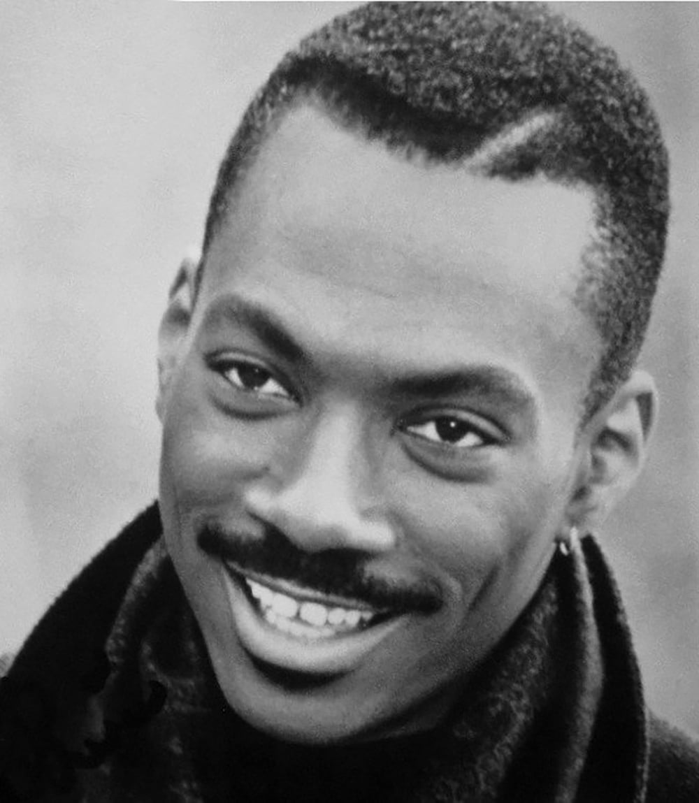 A black and white close-up photo of a man smiling, with short, neatly cut hair, a thin mustache, and an earring in his left ear, wearing a dark jacket or sweater.