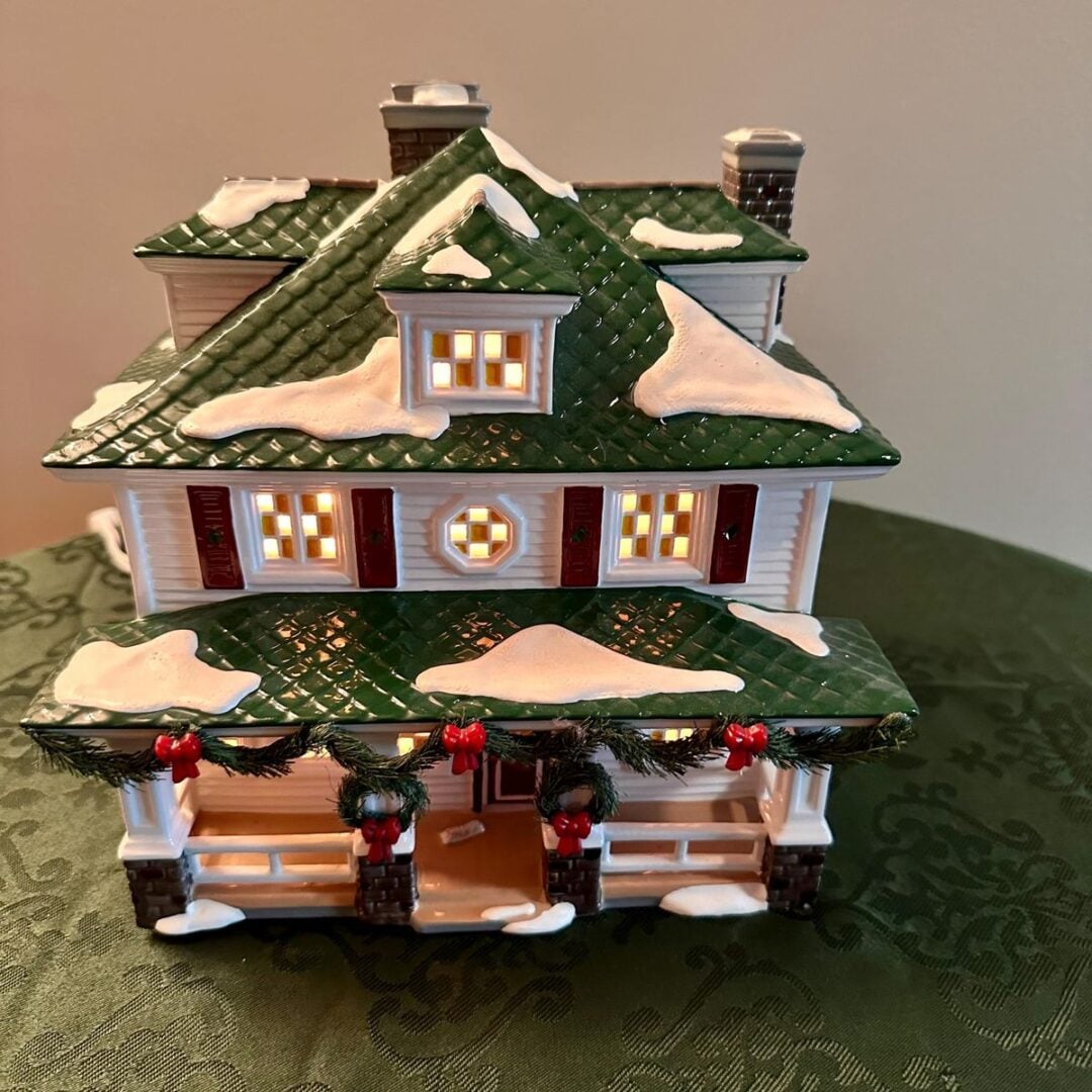 A miniature house decorated for Christmas sits on a green cloth. The house has a green roof with patches of snow, glowing windows, red bows, and garlands hanging on the porch railing.