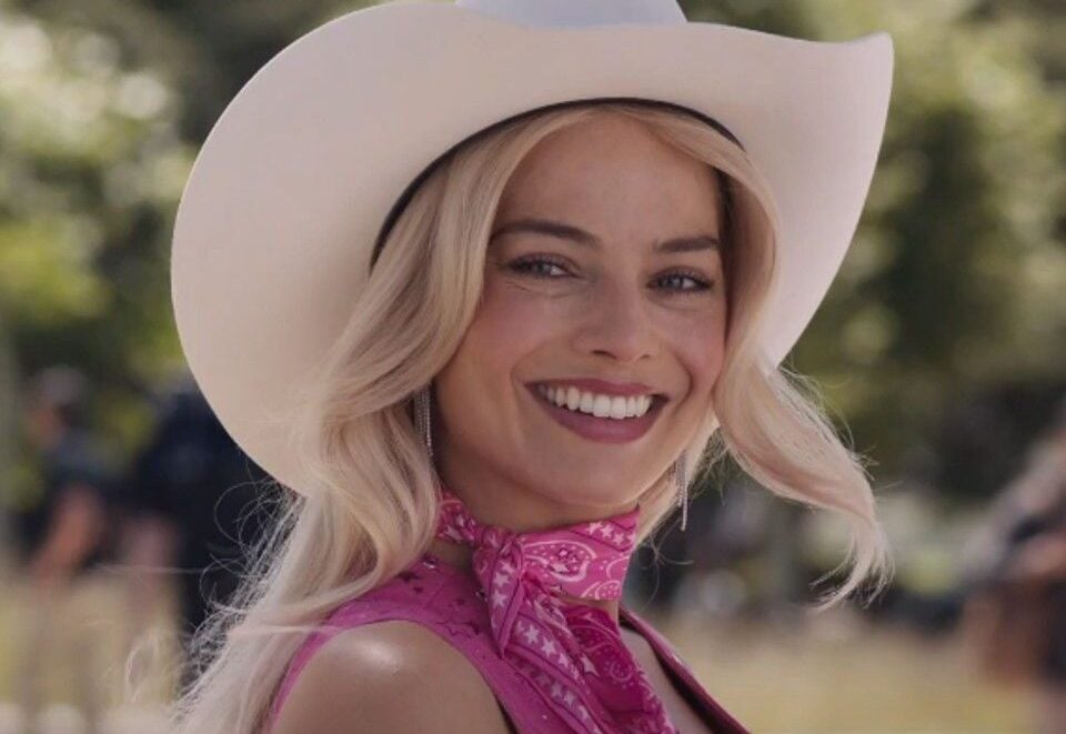 A smiling woman with long blonde hair wears a white cowboy hat, a pink bandana around her neck, and a pink outfit, standing outdoors with greenery in the blurred background.