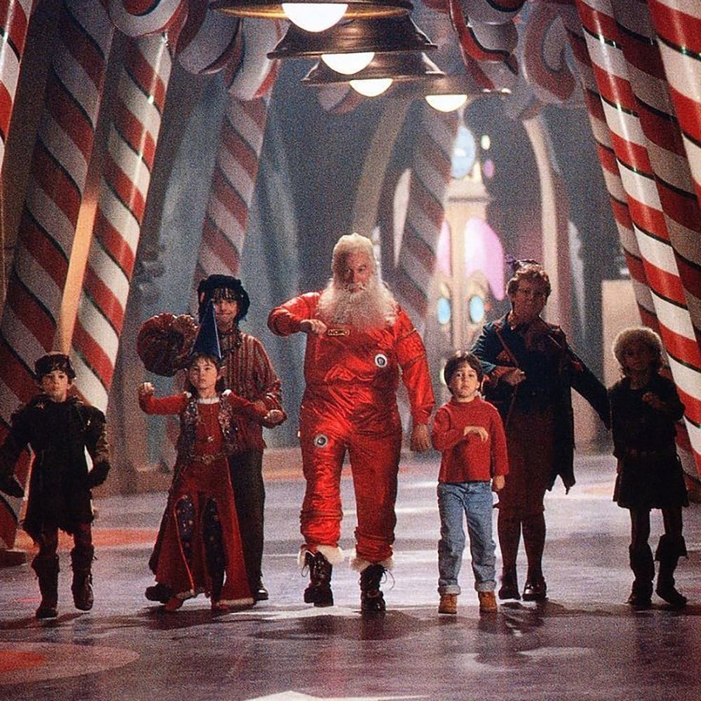 Santa Claus in a red suit skates with five children and two elves in a whimsical hallway decorated with large red and white candy cane columns. The group looks cheerful as they move forward together.