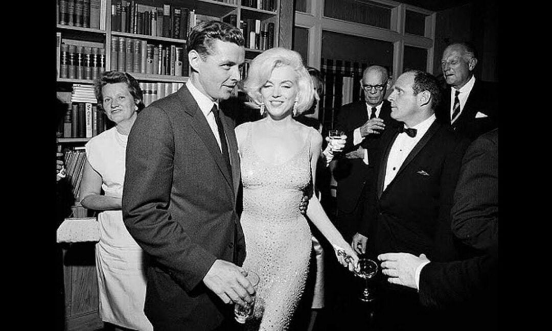 A glamorous woman in a sparkling dress smiles and poses with a suited man at a formal gathering, surrounded by elegantly dressed people holding drinks in a room lined with bookshelves.