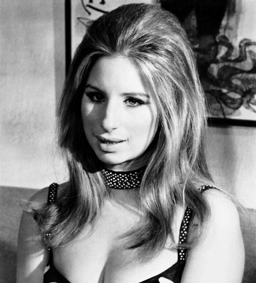 A woman with voluminous, light-colored hair wears a studded choker and a low-cut, decorative dress. She looks slightly past the camera with a neutral expression, seated indoors with framed art visible in the background.