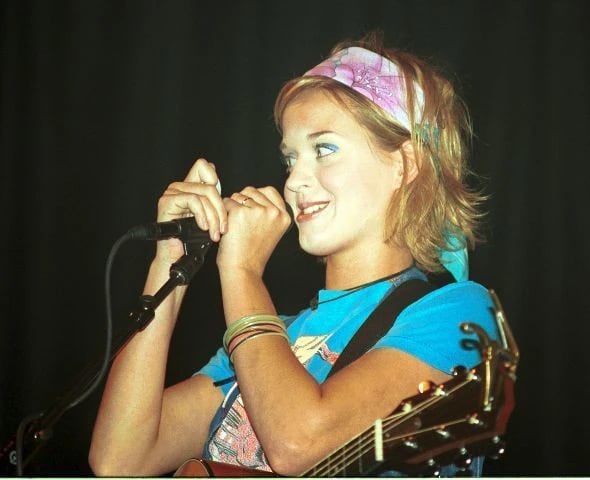 A young woman with short blond hair and a colorful headband smiles while holding a microphone on stage. She wears a blue shirt, several bangles, and a guitar strap over her shoulder, with a dark background behind her.