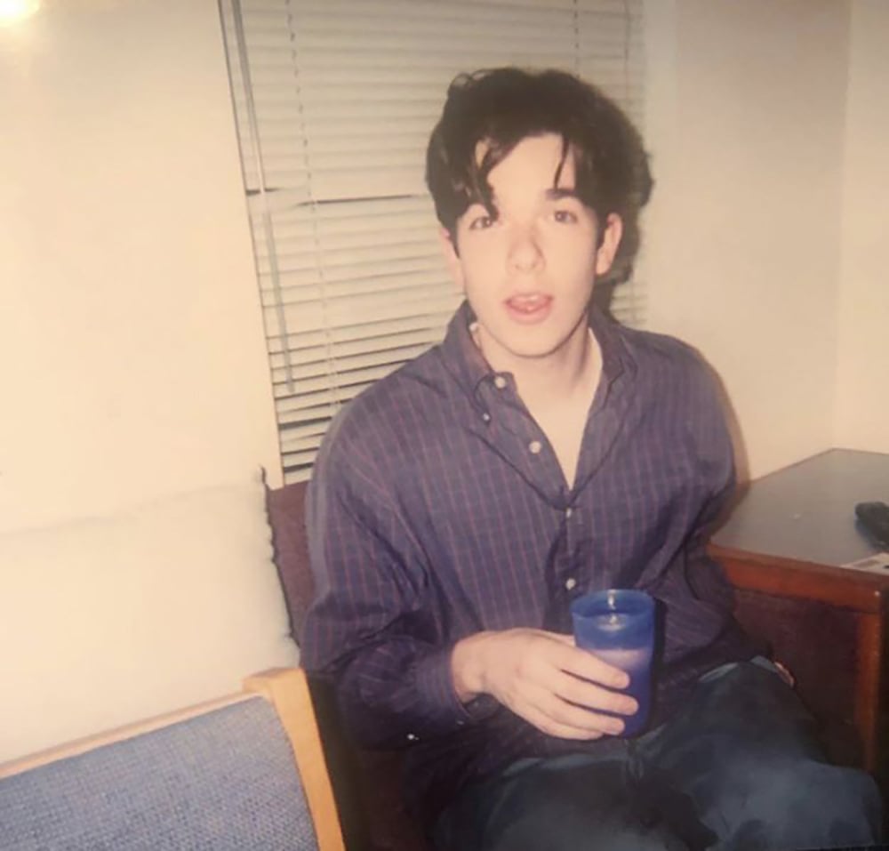 A young man with tousled dark hair sits on a couch holding a blue plastic cup, wearing a striped button-up shirt and looking toward the camera. Blinds cover the window behind him.