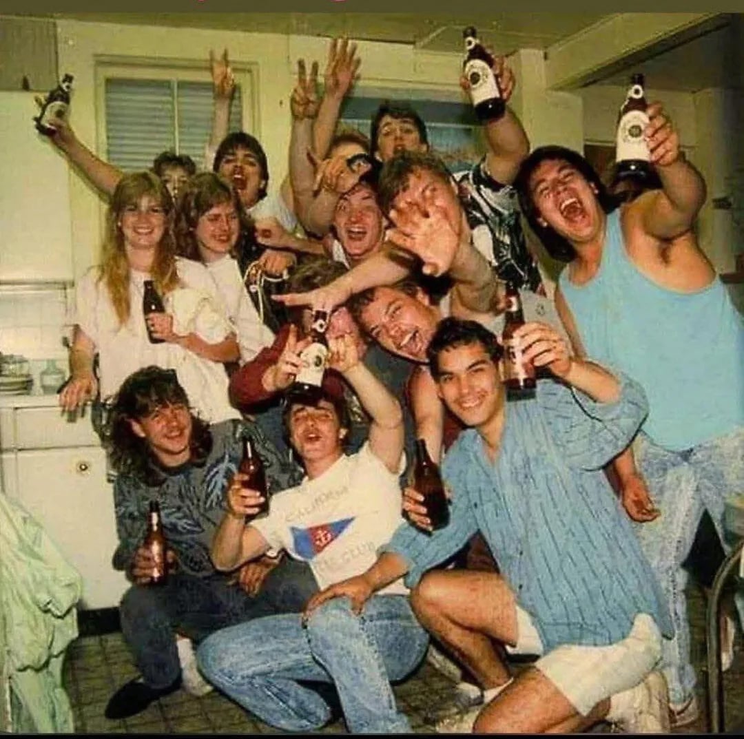 A group of young people in a kitchen, smiling and holding beer bottles, with several making peace signs and excited gestures, creating a lively and celebratory atmosphere.