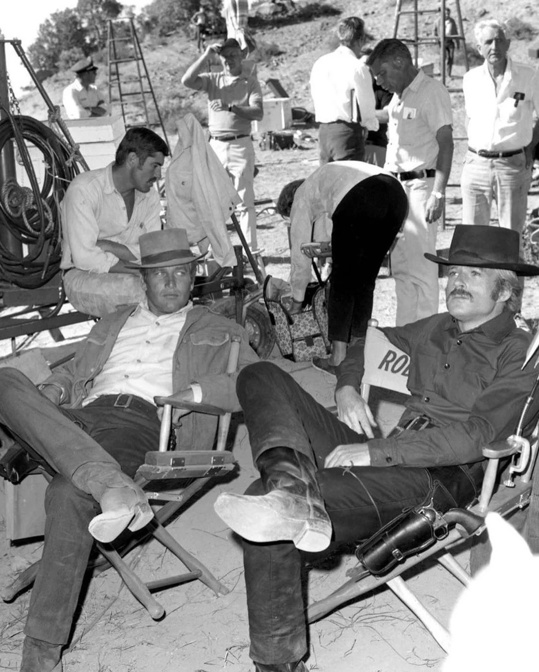 Two men in Western attire relax in chairs on a film set, while several crew members stand and work in the background amid equipment and props under bright outdoor lighting.