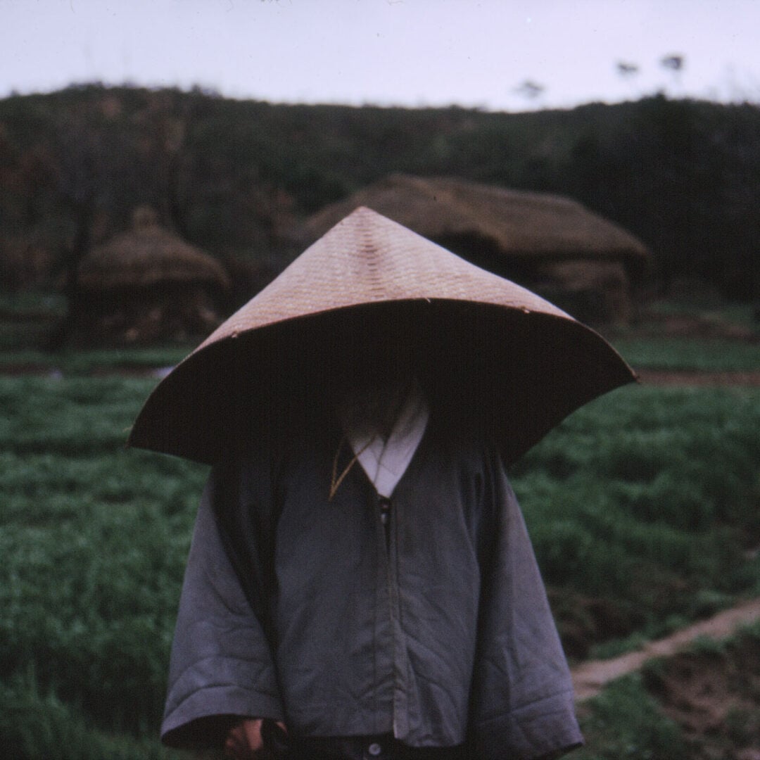 A person wearing a large conical hat and a gray robe stands in a green field with traditional thatched-roof huts in the blurred background. The person's face is obscured by the hat.
