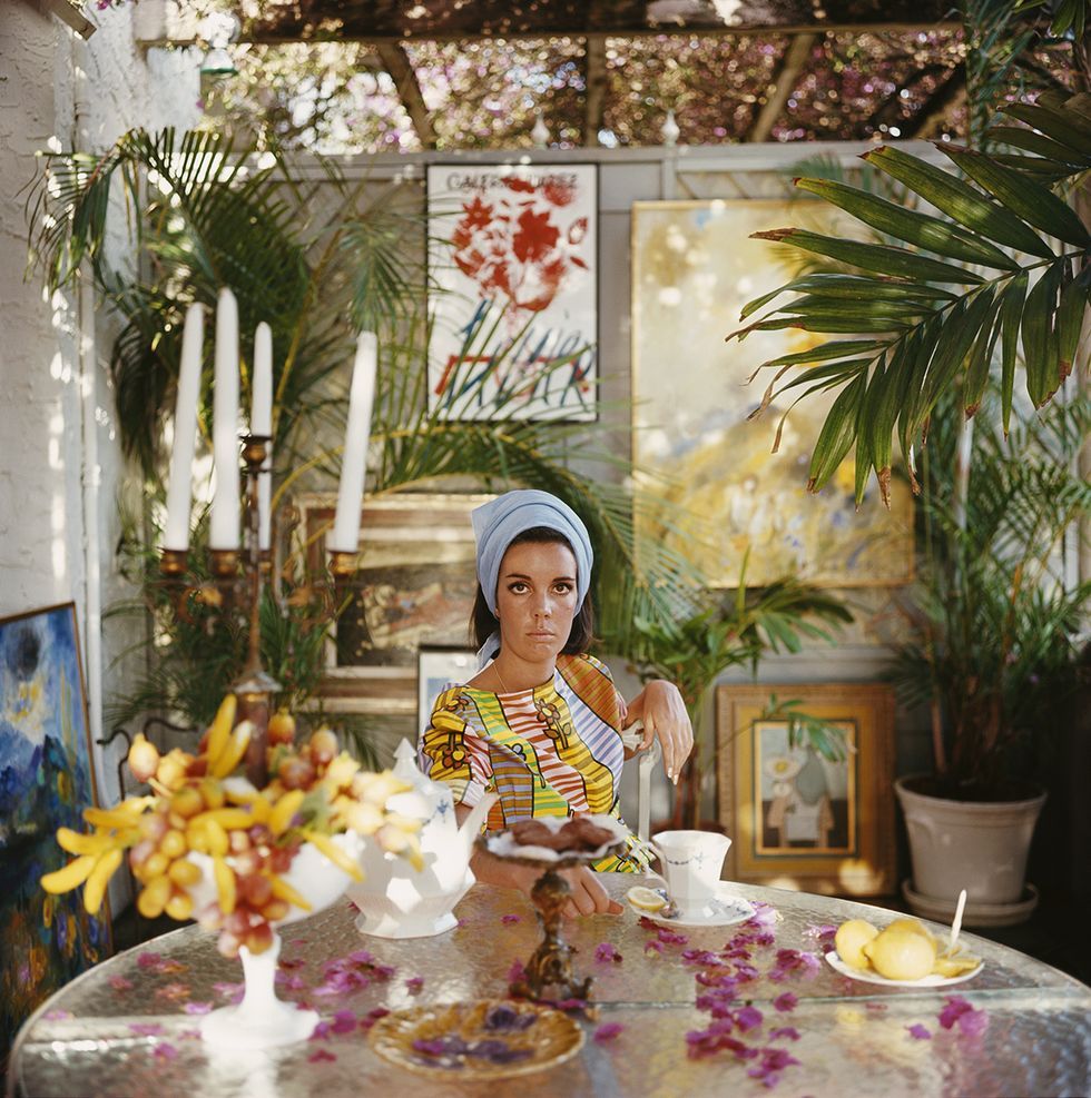 A woman with a blue headscarf sits at a table with fruit, teacups, and flowers, surrounded by lush plants and colorful paintings in a sunlit room.