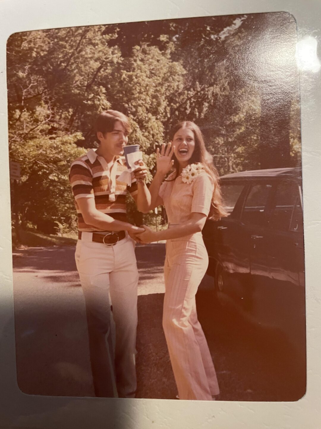 A young man and woman stand smiling outdoors by a car, dressed in 1970s-style clothing. The woman wears a light jumpsuit and a corsage; the man wears striped shirt and light pants, holding a drink. Trees fill the background.