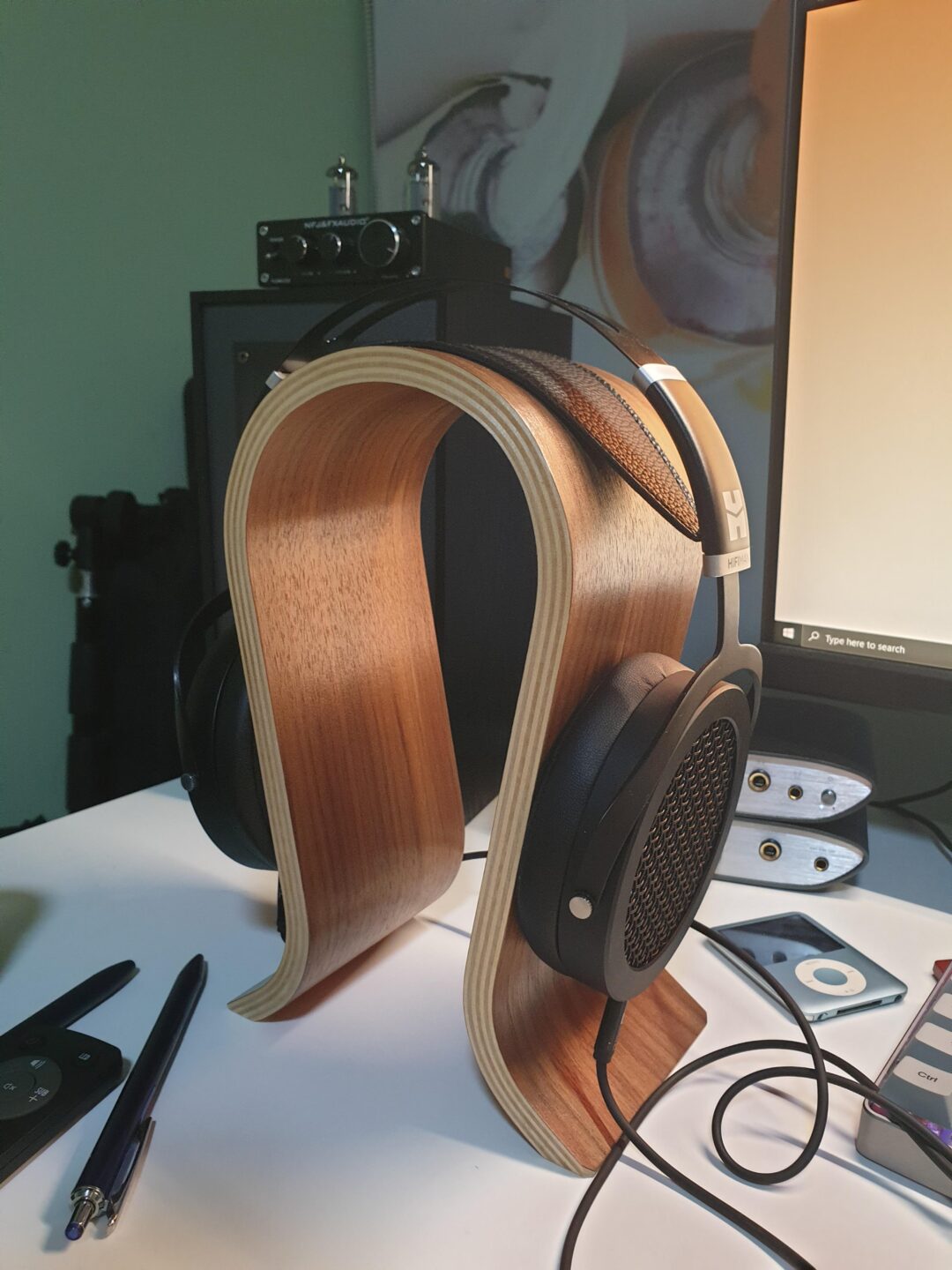 A pair of black open-back headphones rests on a curved wooden headphone stand on a desk, near a monitor and various audio equipment. Pens, a phone, and an audio player are also visible.