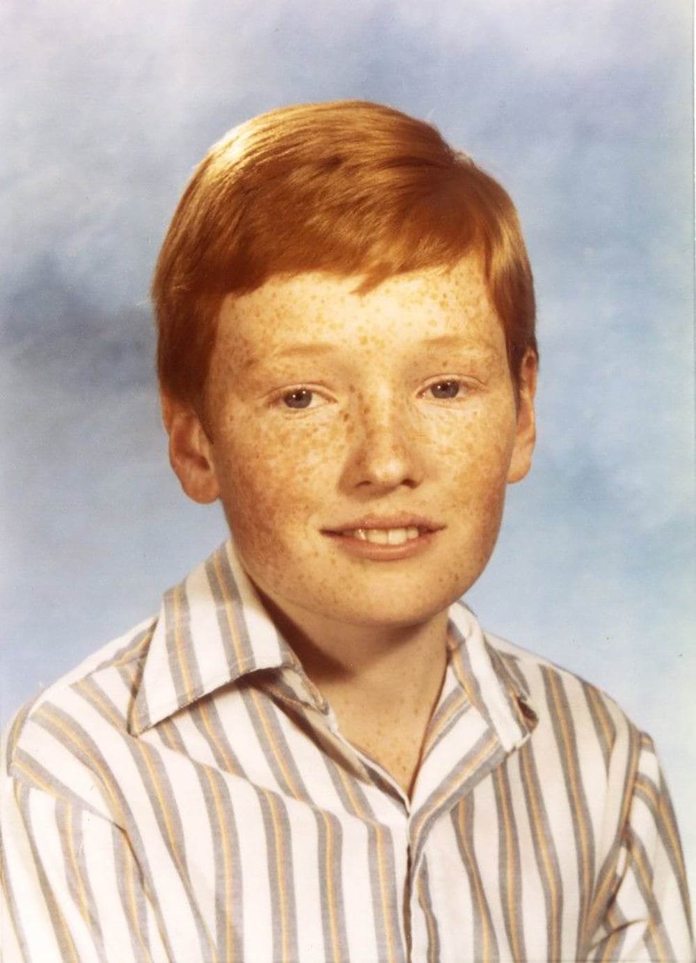 A young boy with red hair and freckles wearing a striped collared shirt poses for a school portrait against a light blue background.