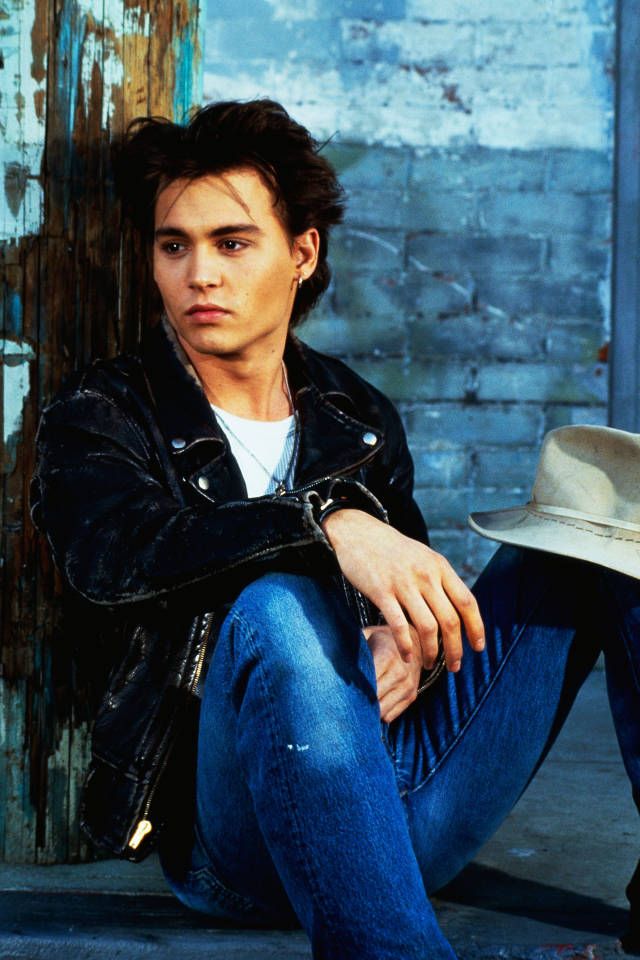 A young man with tousled dark hair wearing a black leather jacket and blue jeans sits on the ground against a weathered wall, holding a cowboy hat and gazing thoughtfully into the distance.
