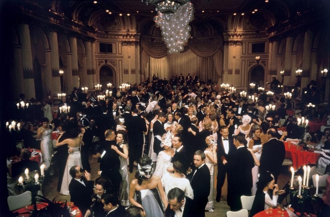 A large, elegant ballroom filled with people dressed in formal attire, dancing and mingling under a grand chandelier, with tables decorated with candelabras surrounding the dance floor.